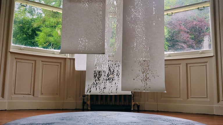 Three rolls of intricately cut white paper hang from the ceiling and float above a dark circular platform in a room.